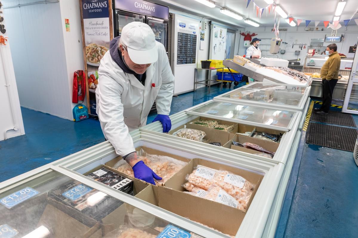 A worker in a white coat arranges frozen seafood in a market display. Another worker and a customer stand nearby amid bright lighting and blue floors.