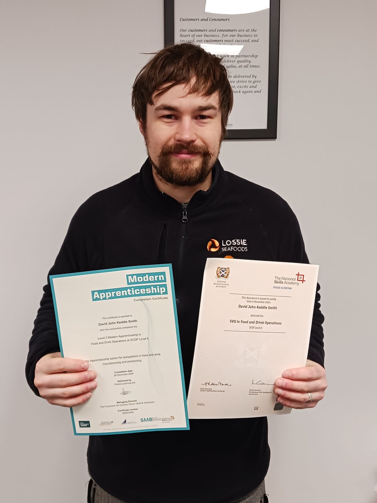 A man with brown hair and a beard holds up two apprenticeship certificates.