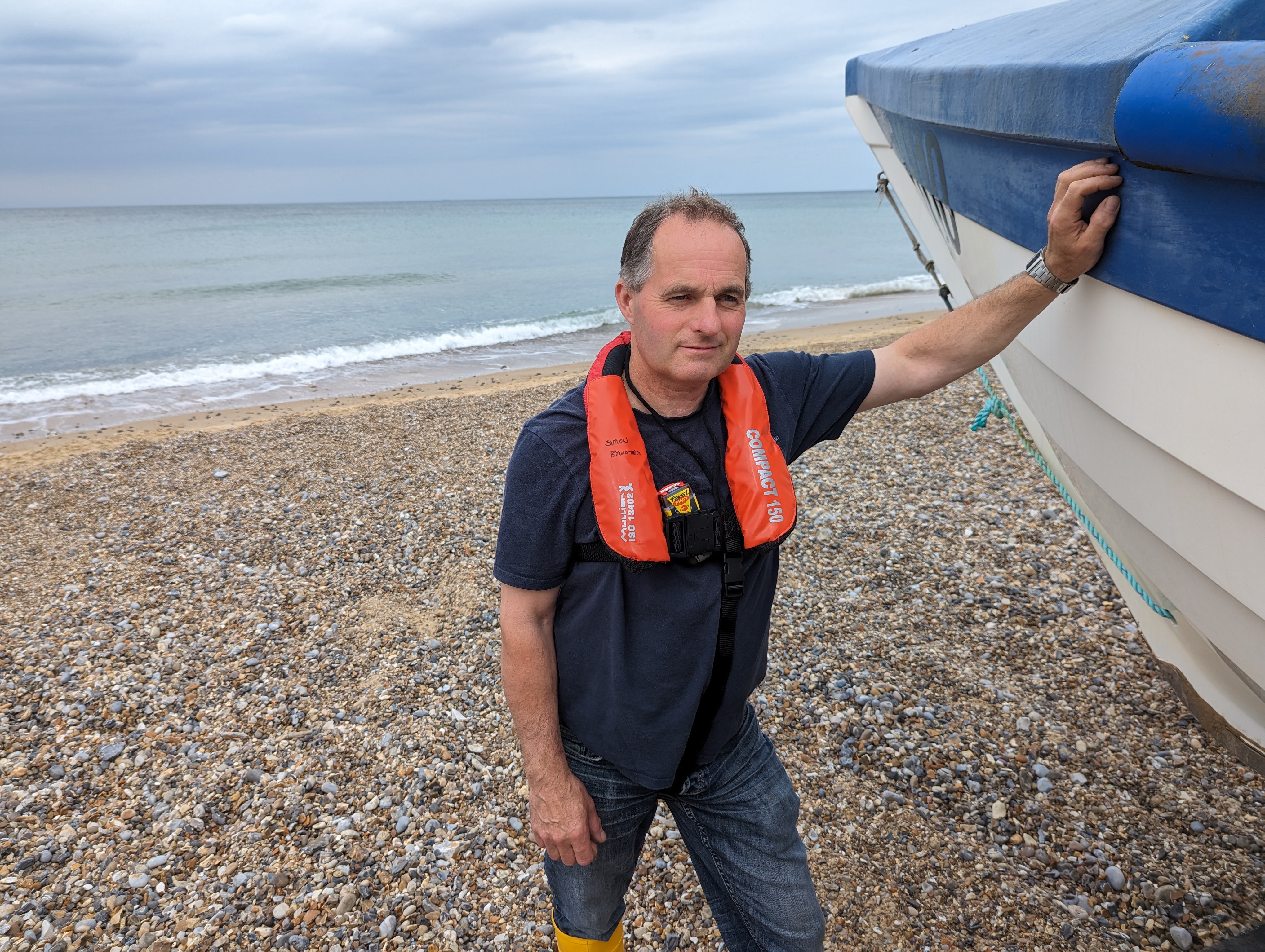 A man wearing a life jacket, looking at the camera, stands on a pebbled beach leaning against a fishing boat.