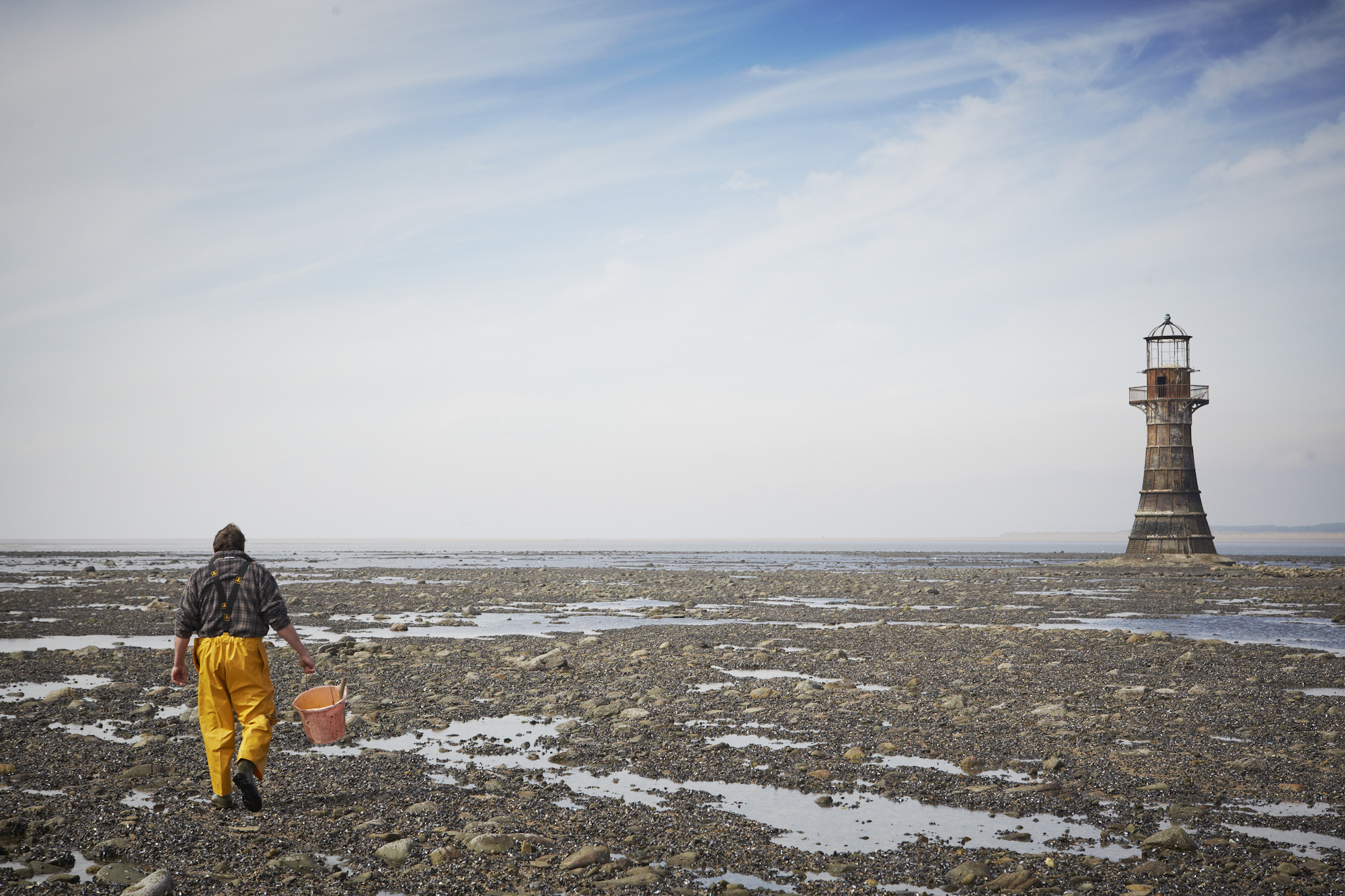 A worker collecting shellfish on a beach with lighthouse in background