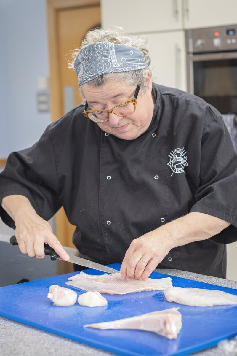 A chef holding a knife prepares thornback ray on a blue chopping board in a kitchen,