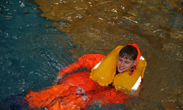 A fisher in the training pool wearing an orange oil skin protective suit and Personal Flotation Device.