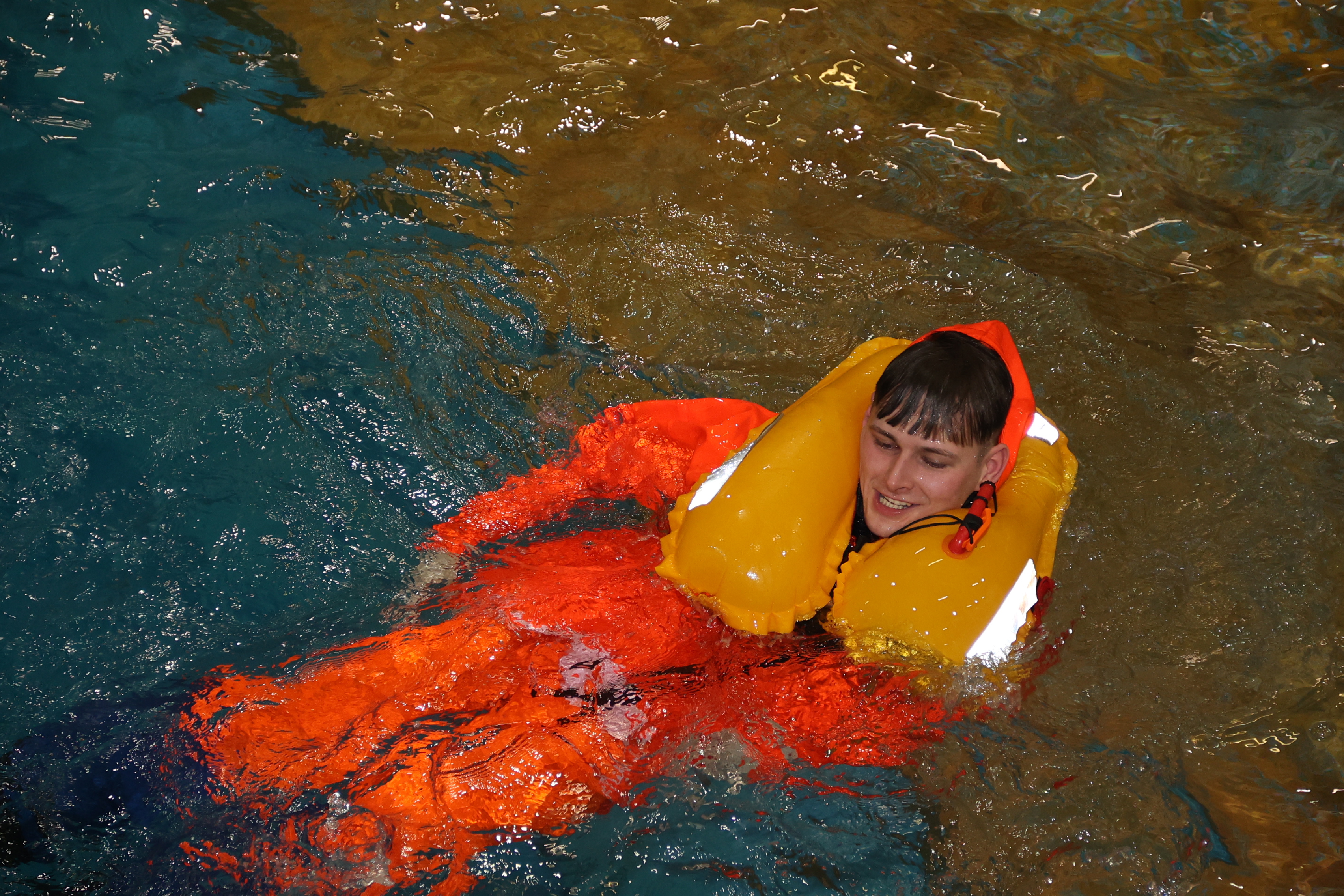 A fisher in the training pool wearing an orange oil skin protective suit and Personal Flotation Device.