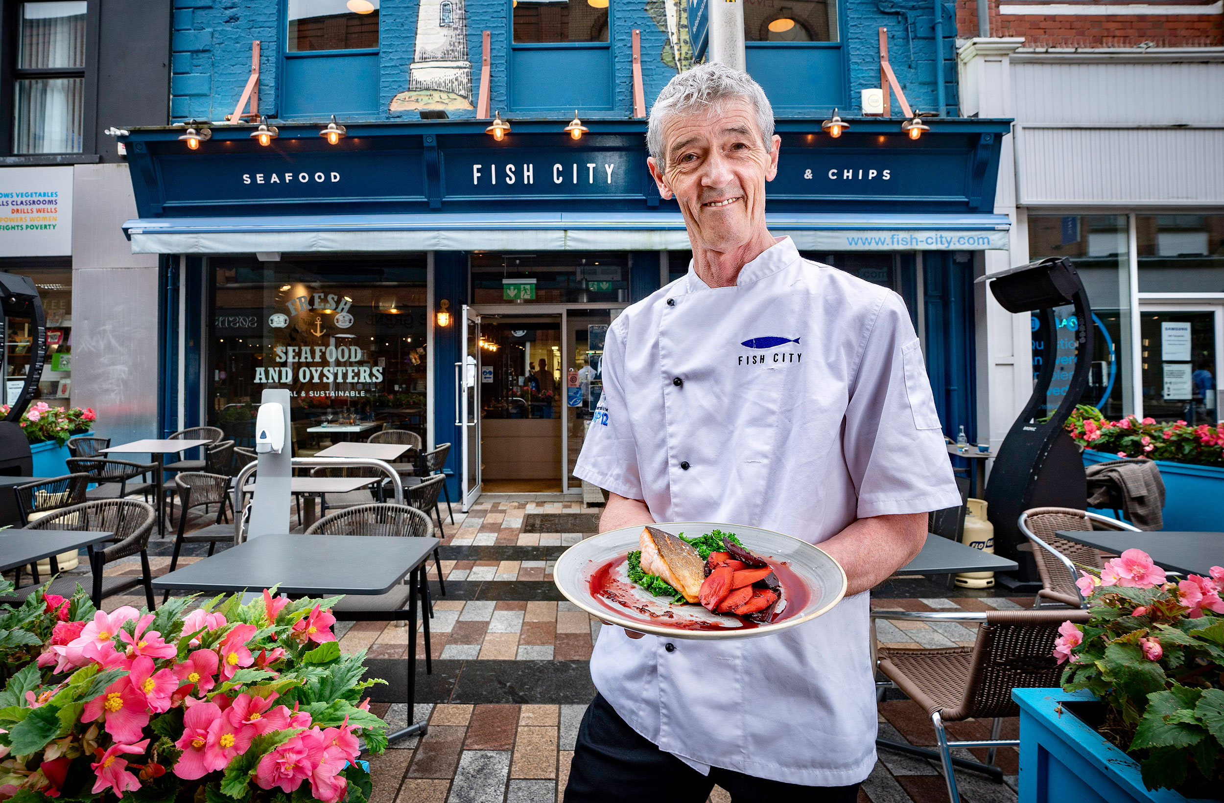 Chef holding a plate of seafood in front of Fish City restaurant, outdoor seating with flowers and brick walkway visible.