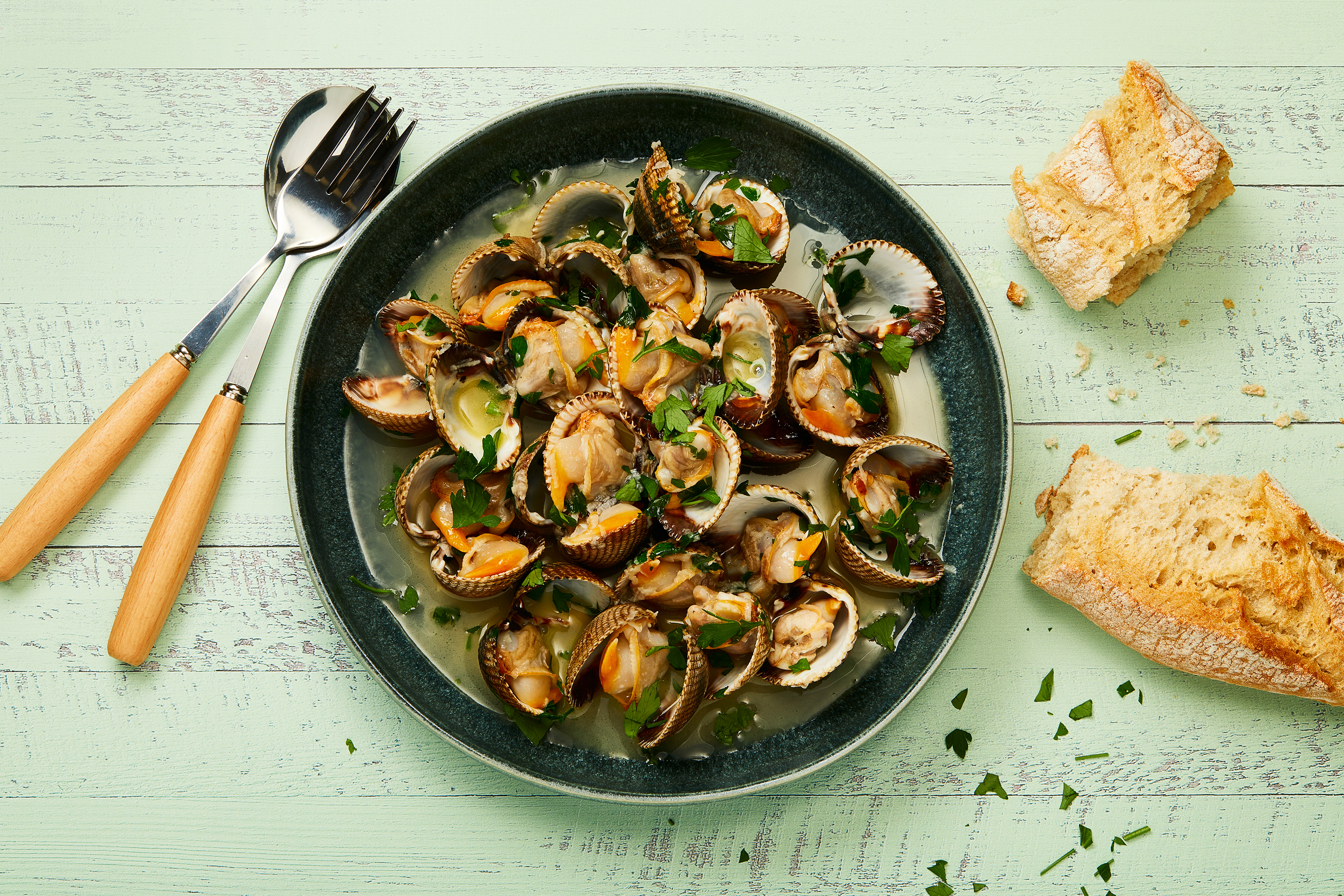 A bowl of steamed clams in a light broth, garnished with parsley, served with crusty bread on a mint green table, alongside wooden-handled utensils.