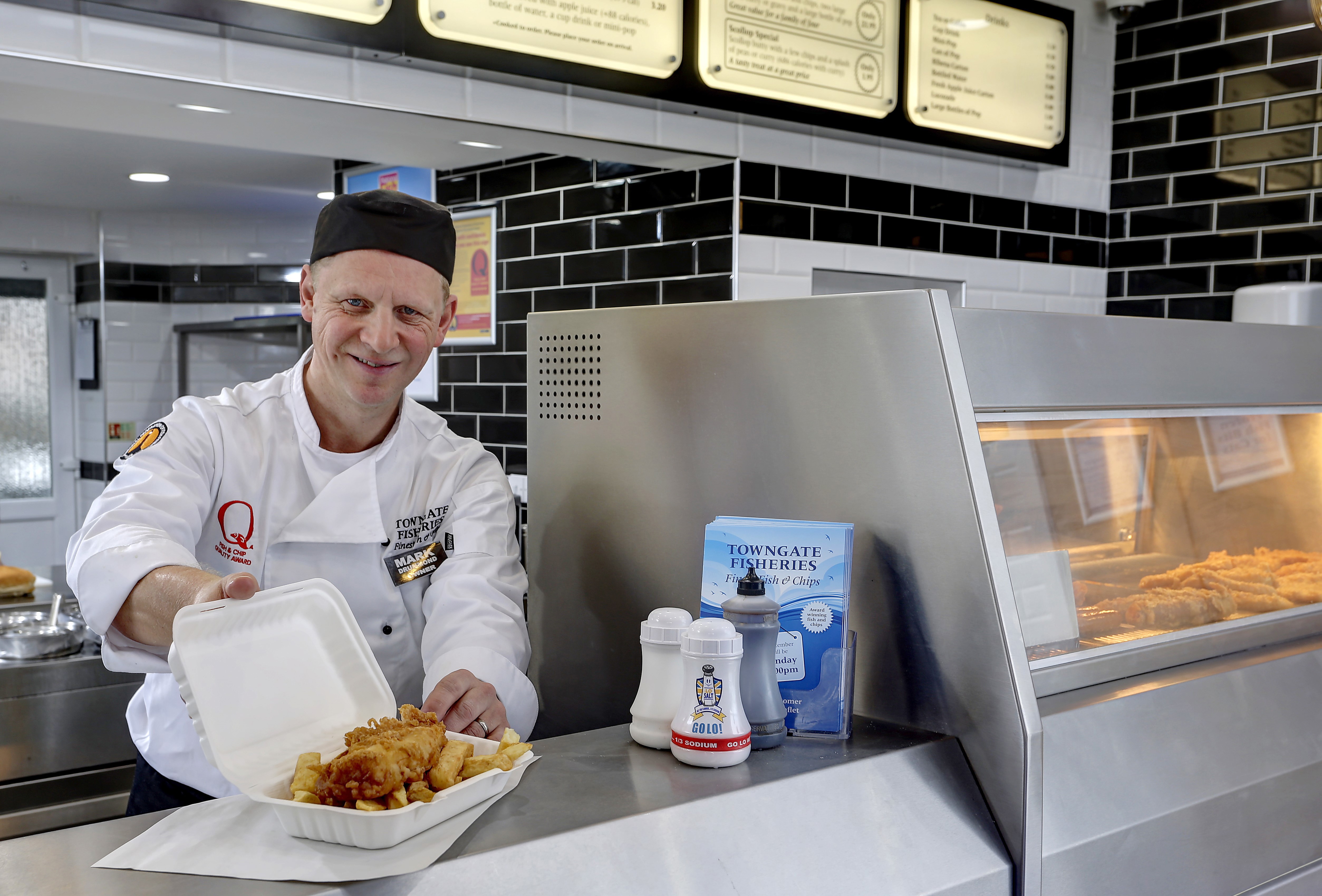 A chef in a black hat and white uniform serves fish and chips in a takeaway box at a fish and chip shop counter.