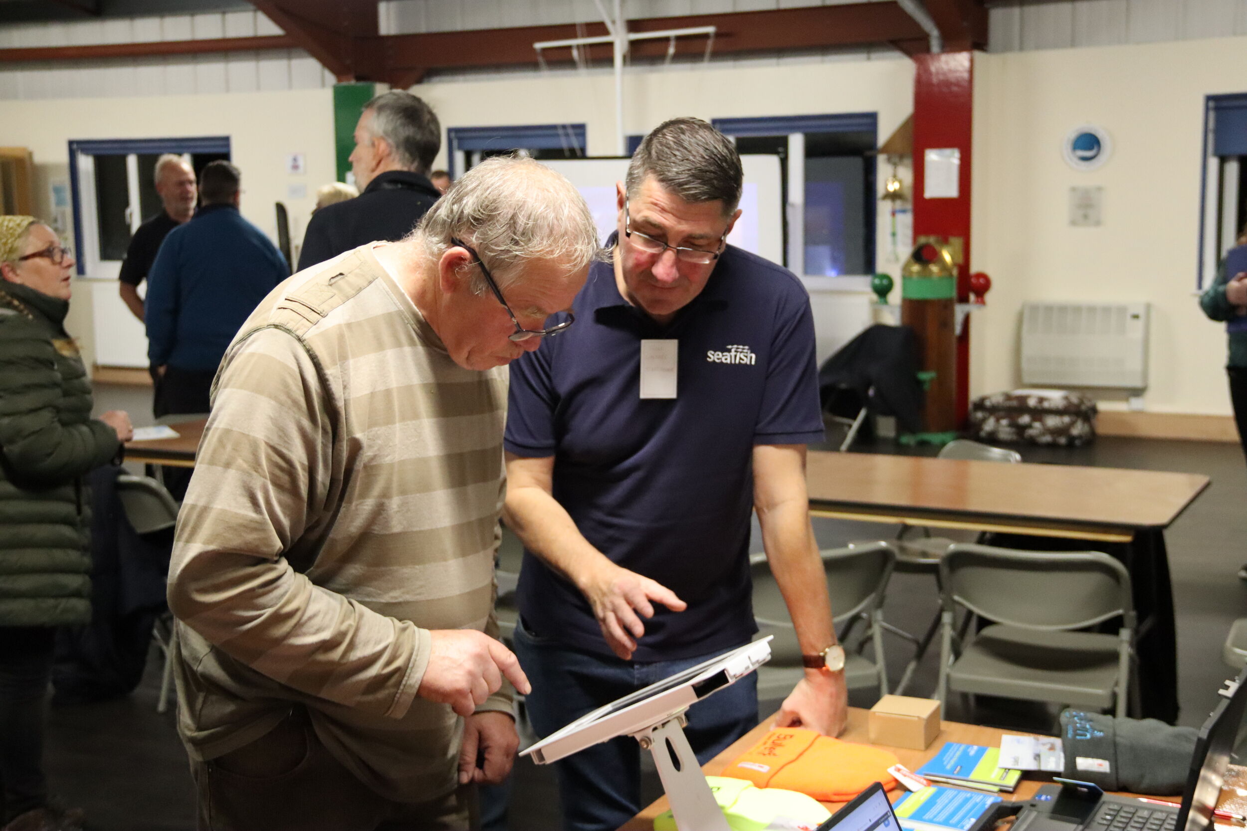 Two men looking at an iPad on a desk with people standing speaking in the background.