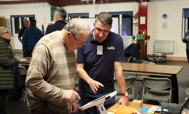 Two men looking at an iPad on a desk with people standing speaking in the background.