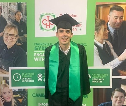 A male stands smiling wearing a black gown, green sash and a square academic cap in front of a banner promoting apprentices.